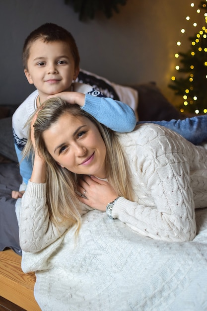 젊고 매력적인 금발 엄마와 그녀의 아들이 크리스마스 거실에서 웃고 껴안고 있습니다.