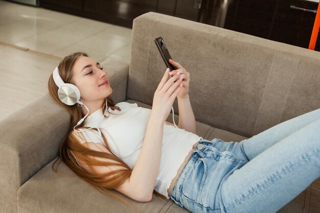 Young attractive blonde woman listens to music and looks at the smartphone screen while lying on sofa in living room