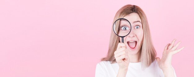 Young attractive blonde woman shocked while looking through a magnifying glass over pink background