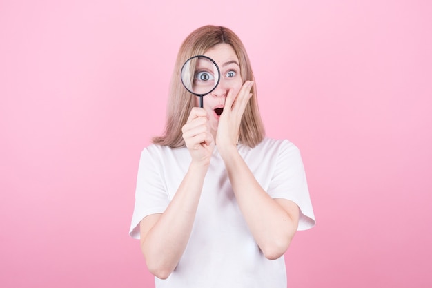 Young attractive blonde woman shocked while looking through a magnifying glass over pink wall