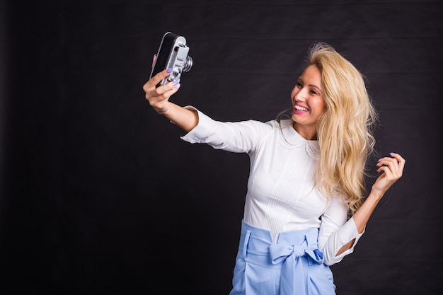 Young attractive blonde woman taking selfie on vintage camera on black surface.