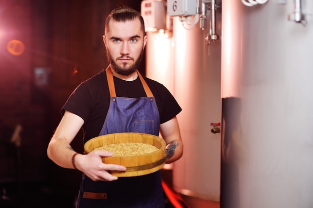 Young attractive brewer enjoys the aroma of malt on the background of the brewery and beer tanks