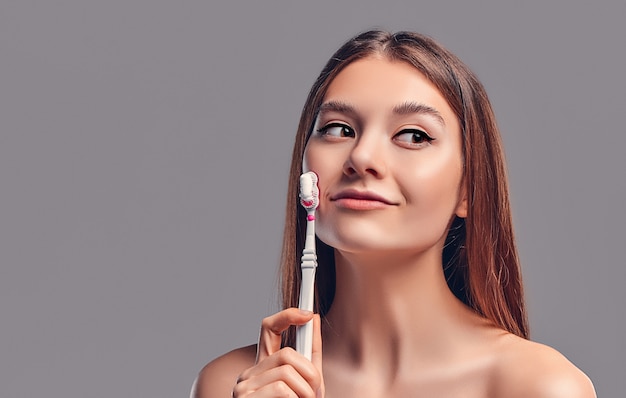 Young attractive brunette girl with loose hair uses a toothbrush and paste isolated on a gray background.