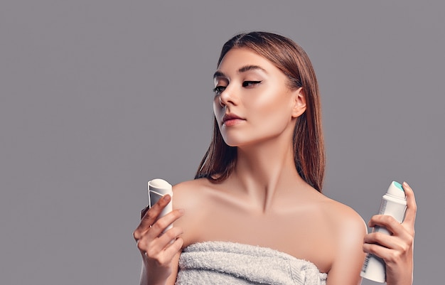 Young attractive brunette girl with loose hair with deodorant stick and antiperspirant aerosol isolated on gray background. Body care.