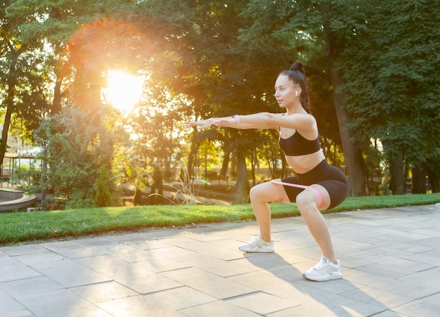 Young attractive brunette woman in sportswear exercises with fitness rubber bands in the park at sunrise