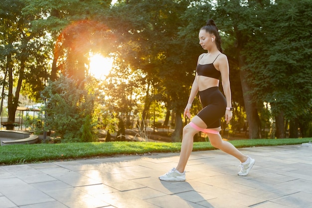 Young attractive brunette woman in sportswear exercises with fitness rubber bands in the park at sunrise