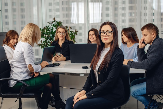 Young attractive brunette woman wearing glasses sitting on a chair in the office surrounded by her colleagues immersed in work. Concept of work