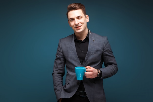 Il giovane uomo d'affari attraente in rivestimento grigio e camicia nera tiene la tazza blu
