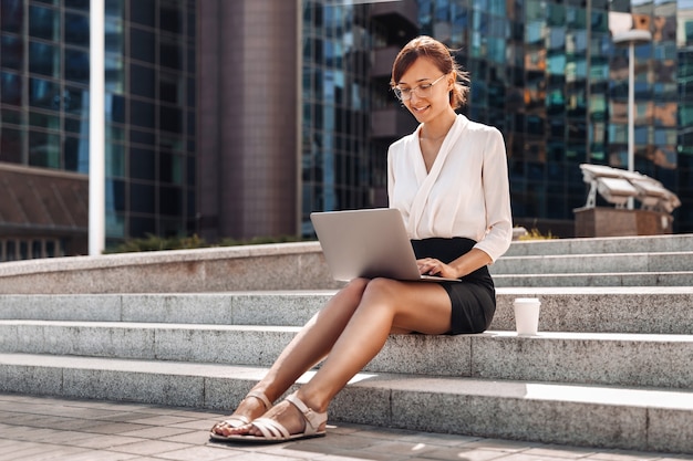 Молодая привлекательная бизнес-леди, работающая за ноутбуком на улице перед бизнес-центром, сидя на ступеньках