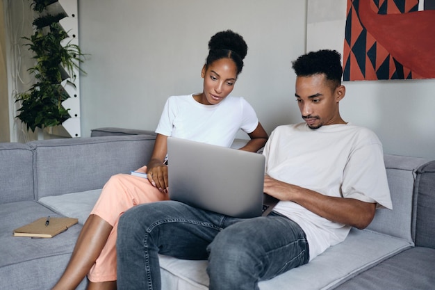 現代の家のソファで一緒にラップトップに思慮深く取り組んでいる若い魅力的なカジュアルなアフリカ系アメリカ人のカップル