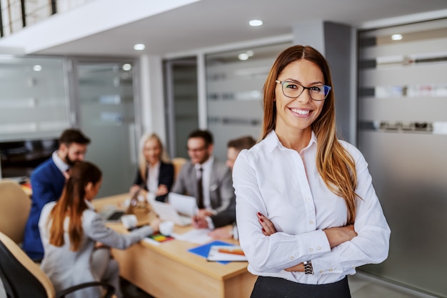 Photo young attractive caucasian businesswoman in formal wear standing in boardroom with hands crossed and looking at camera. in background are her colleagues working on project.