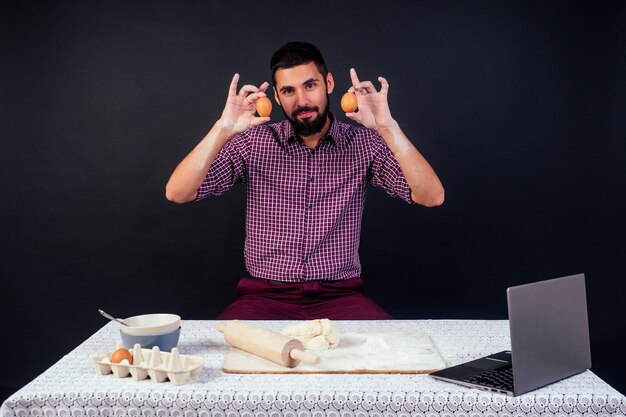 Young and attractive caucasian man baker with beard makes the dough sprinkled with flour on a black background in the studio . Confectioner bakes cake