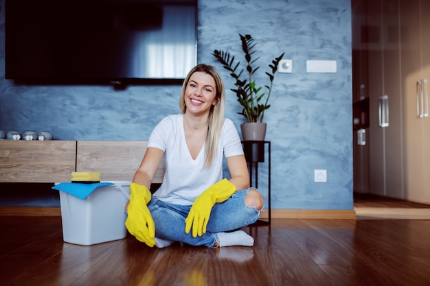 Young attractive caucasian smiling blonde housewife with rubber gloves on hands sitting on the floor with legs crossed and looking. Next to her is bucket.