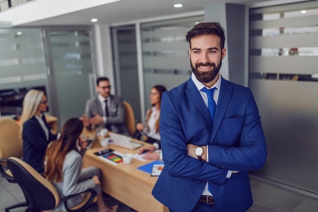 Young attractive Caucasian smiling satisfied businessman in suit standing in boardroom with arms crossed