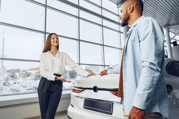 Young attractive caucasian woman salesperson in car showroom showing a car to her male African American client