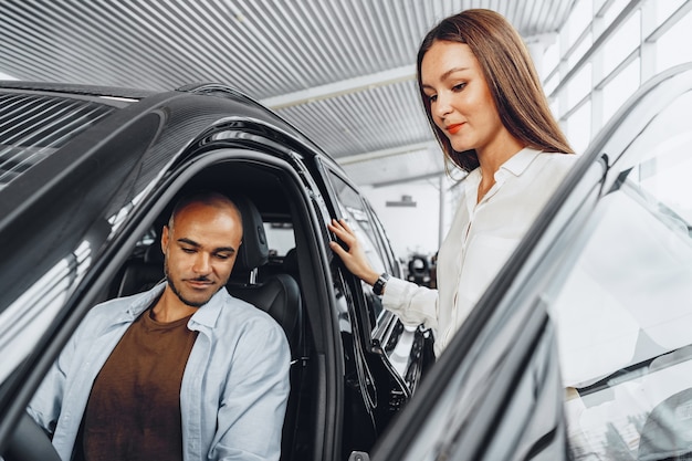 彼女の男性のアフリカ系アメリカ人のクライアントに車を見せて車のショールームで若い魅力的な白人女性の販売員