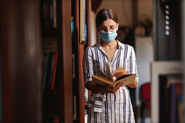 Young attractive college girl with face mask on standing in library and browsing material in a book for school project during corona virus pandemic.