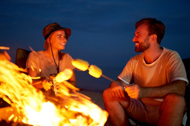 Young attractive couple sit on folding chairs near the tent and\
grill corn on a fire and have fun talking at night near the\
sea.
