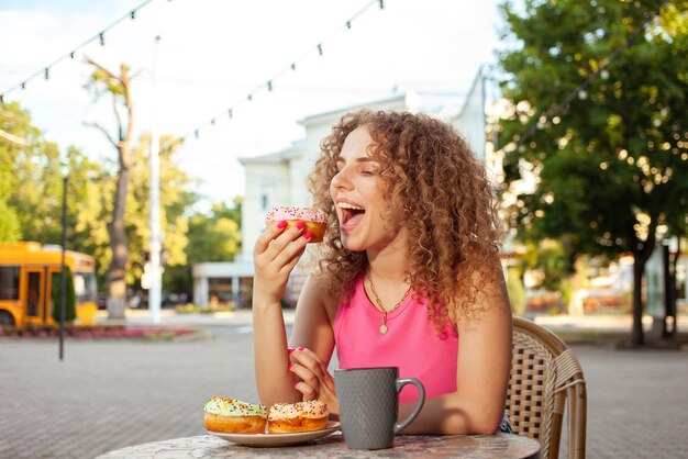 Photo young attractive curly woman sits at table in cafe on summer terrace colorful delicious donuts