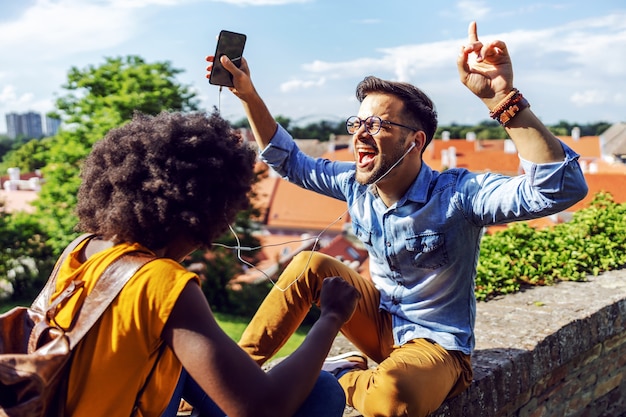 Young attractive cute multicultural hipster couple enjoying music while sitting on the wall.