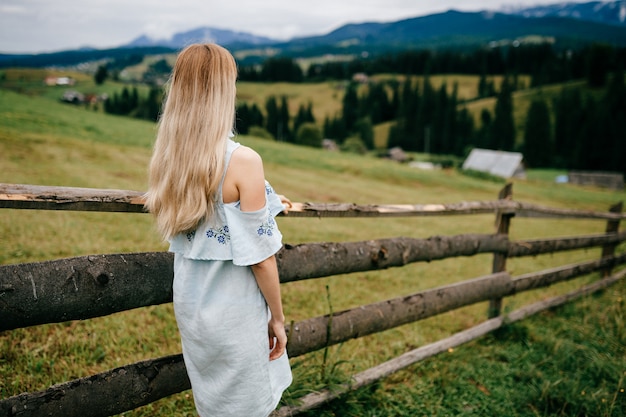 Молодая привлекательная элегантная блондинка в синем платье позирует возле забора в сельской местности