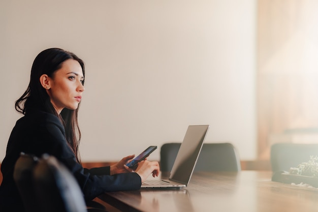 Young attractive emotional girl in business-style clothes sitting at a desk on a laptop and phone