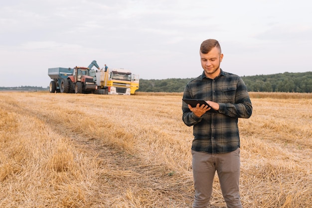 Giovane agricoltore attraente con laptop in piedi nel campo di grano con mietitrebbia in background