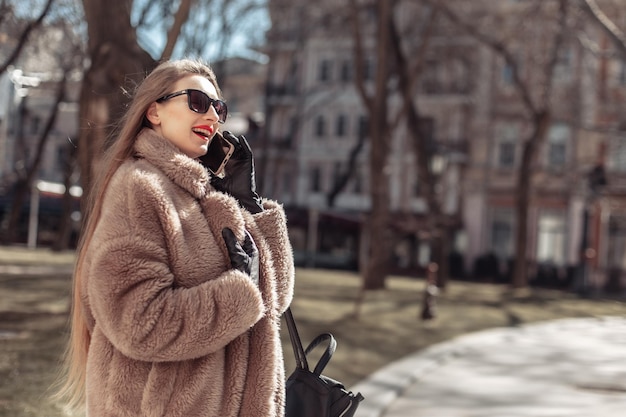 Young attractive fashion blonde woman dressed in fur coat and sunglasses talking on the phone in city