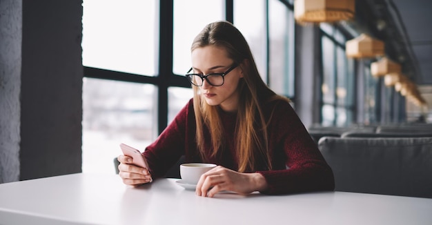 お気に入りのカフェに座ってスマートフォンで友達とおしゃべりする若い魅力的な女子学生