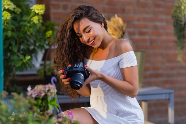 Young attractive female student or tourist using a mirrorless camera while walking in summer city wo...