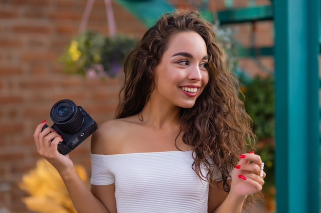 Young attractive female student or tourist using a mirrorless camera while walking in summer city wo...