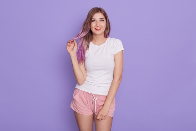 Young attractive female with flirting expression wearing casual style attire, looking directly at camera, pulling her curl aside and smiling, isolated over lilac background.