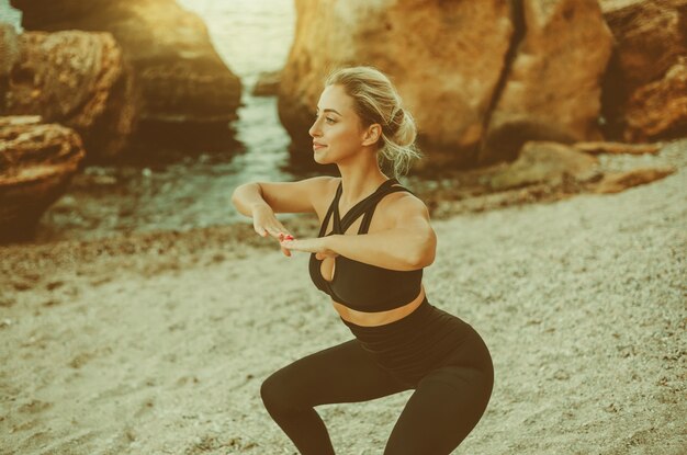 Young attractive fitness woman in sportswear doing squats in the sand at wild beach. Sport training. Workout outdoors