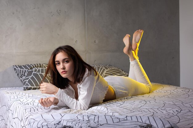 A young attractive girl dressed in white pajamas is resting on a bed in a bedroom in the evening lighting