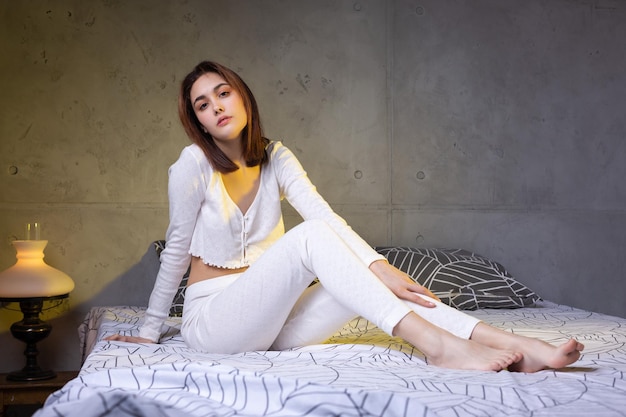 A young attractive girl dressed in white pajamas is resting on a bed in a bedroom in the evening lighting