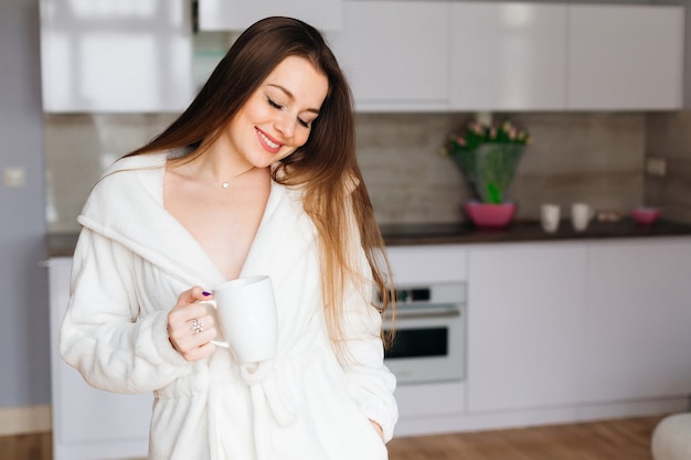 Молодая привлекательная девушка, пить утренний кофе из большой белой чашки, улыбаясь, утренний портрет. Скопируйте пространство.