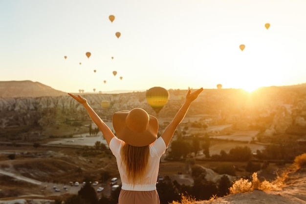 Young attractive girl stands on the mountain with flying air balloons on the background Cappadocia