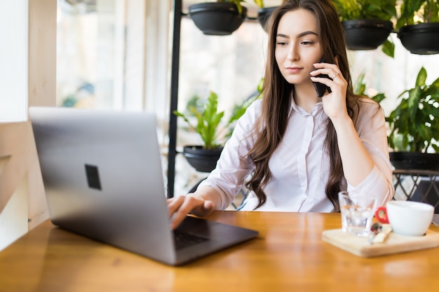 Young attractive girl talking on mobile phone and smiling while sitting alone in coffee shop during free time and working on tablet computer. Happy female having rest in cafe. Lifestyle