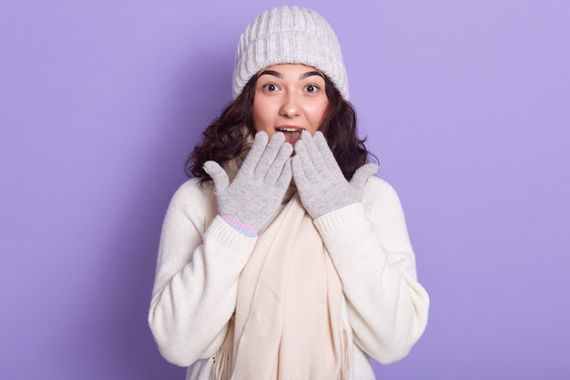 Young attractive girl wearing white sweater, warm cap, scarf and gloves, having light makeup, covering her mouth with her hands