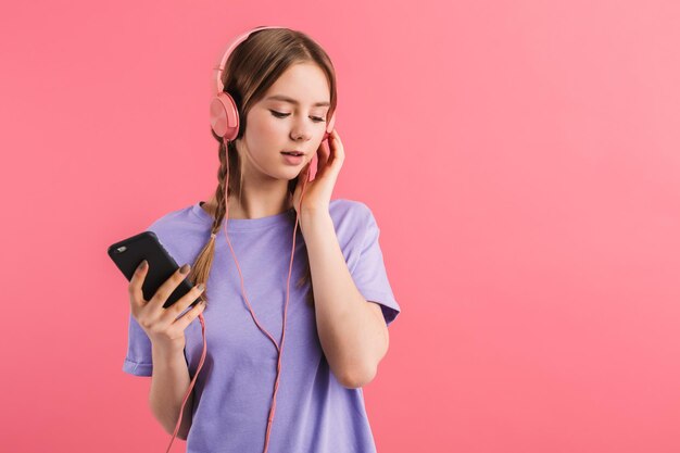 Young attractive girl with two braids in lilac t shirt listening music in headphones with cellphone in hand while dreamily looking aside over pink background