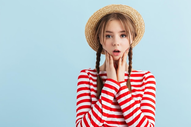 Young attractive girl with two braids in straw hat and red striped vest holding hands near face while amazedly looking in camera over blue background