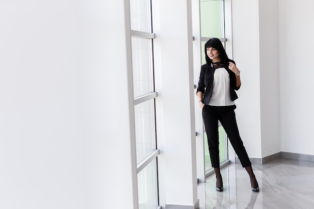 Young Attractive happy brunette woman with long hair standing near the window in office, smiling, looking out to window.