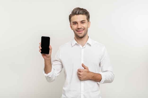 Foto giovane uomo attraente e felice che indossa una camicia bianca che tiene in mano uno smartphone con schermo vuoto e mostra il pollice in su