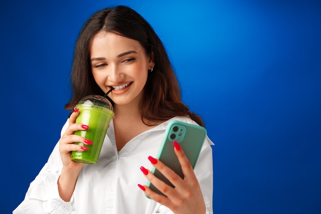 Young attractive happy woman holding a cup of green smoothie and using her smartphone against blue b...