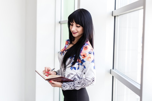 Young Attractive happy woman standing near the window, writing in a notebook, looking to notebook, smiling.