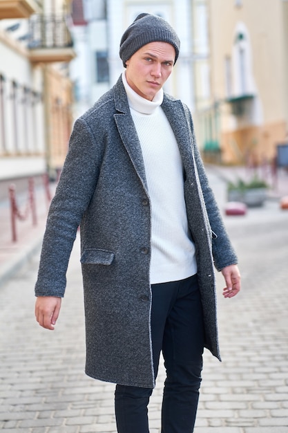 写真 通りを歩いて灰色のコート、白いセーター、黒のジーンズを身に着けている若い魅力的な流行に敏感な男