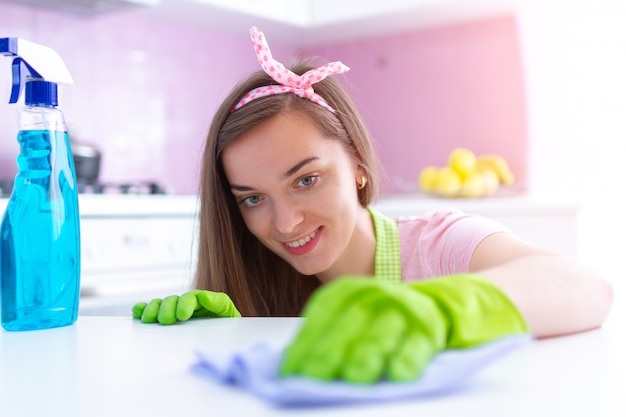 Photo young attractive housewife in protective rubber gloves wiping dust off furniture at kitchen at home using a rag and spray. spring cleaning and housekeeping
