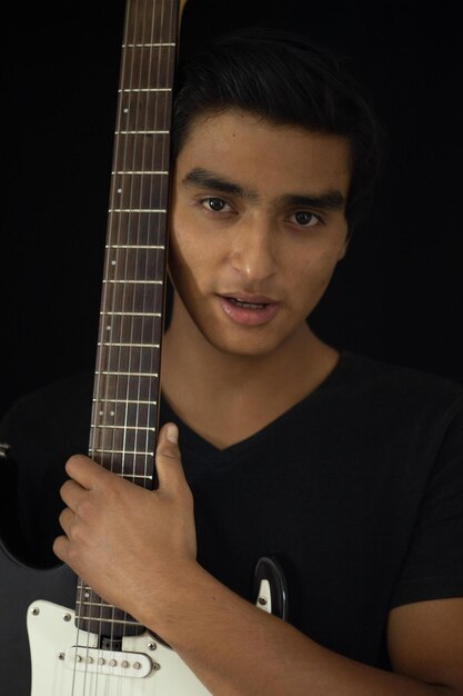 Young and attractive latin musician posing inside a studio and hugging his electric guitar and looking at the camera.