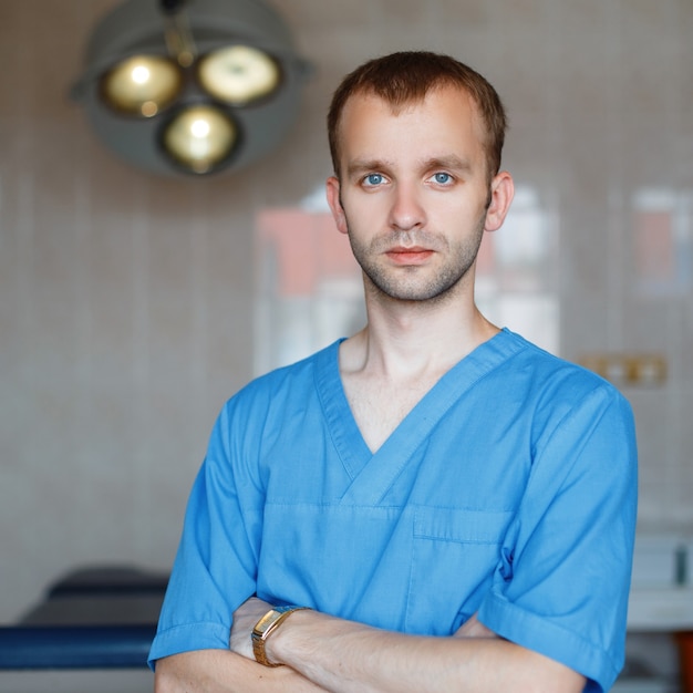 Young attractive male doctor in blue medical clothing is standing in the hospital in the operating room