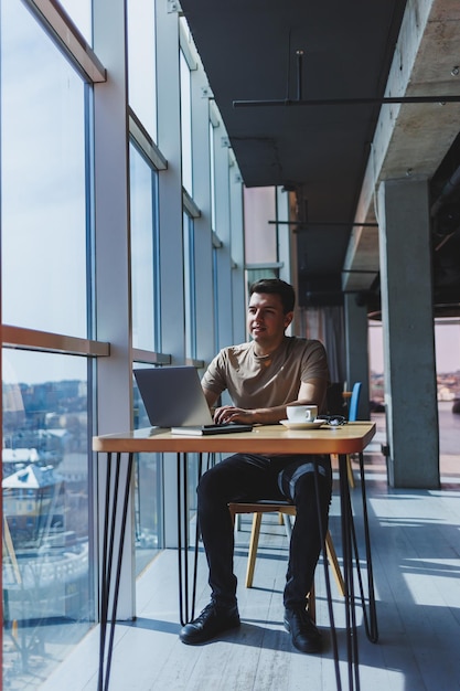 Un giovane freelance maschio attraente si siede all'interno di una caffetteria e guarda fuori dalla finestra blogger bello che scrive idee sul computer portatile mentre è seduto al tavolo di legno nella caffetteria con il computer portatile